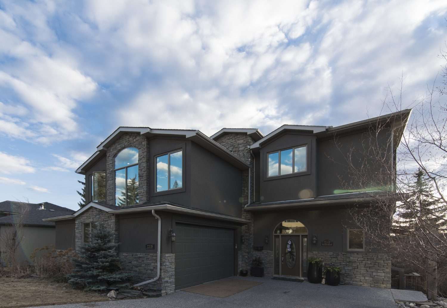 3464 Two-story house with dark siding, large windows, and a stone facade. Features a two-car garage, potted plants by the entrance, and a clear sky with scattered clouds.