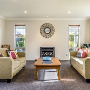 Living room with two beige sofas, a wooden coffee table, a round wall mirror, and a small fireplace. Large windows on either side allow natural light to illuminate the space.