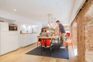 Three people are gathered around a dining table in a modern kitchen with exposed brick walls and contemporary lighting.