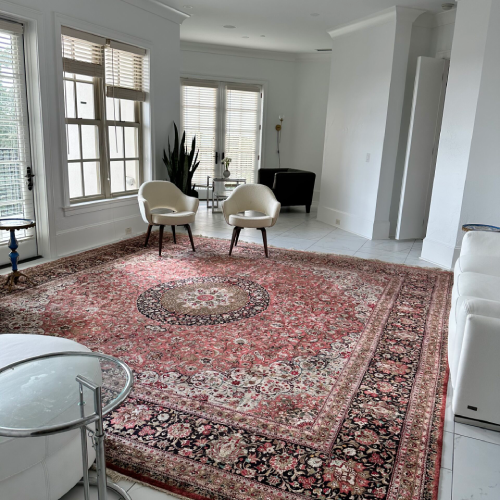 A spacious living room with a large patterned red rug, two white chairs, a plant by the window, and white walls with natural light streaming in.