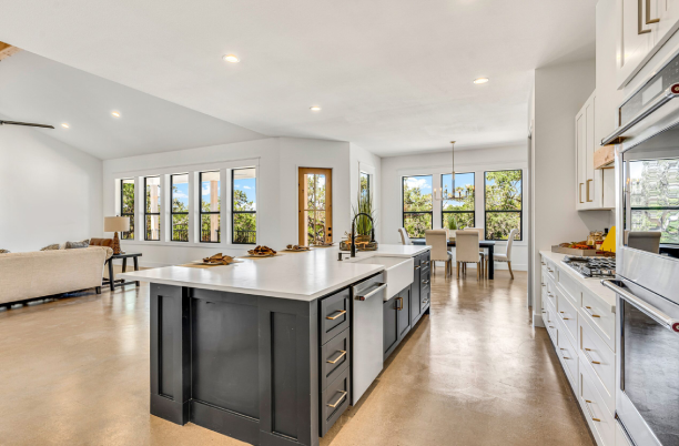 Modern open-concept kitchen with a large island, white cabinets, and stainless steel appliances, leading to a dining area and living room with large windows.