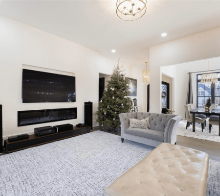 Modern living room with a large TV, fireplace, grey sofa, ottoman, and a decorated Christmas tree; dining area visible in the background.