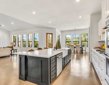Modern open-concept kitchen with a large island, white cabinets, stainless steel appliances, and adjacent dining and living areas with large windows.