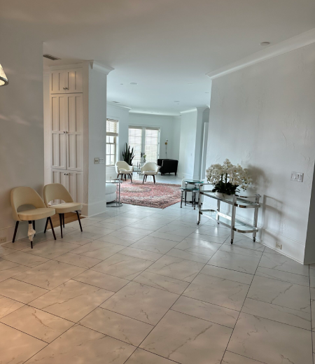 Bright, modern living space with white tiled floors, two beige chairs, a glass console table with white flowers, and a sitting area with chairs and a rug near large windows.